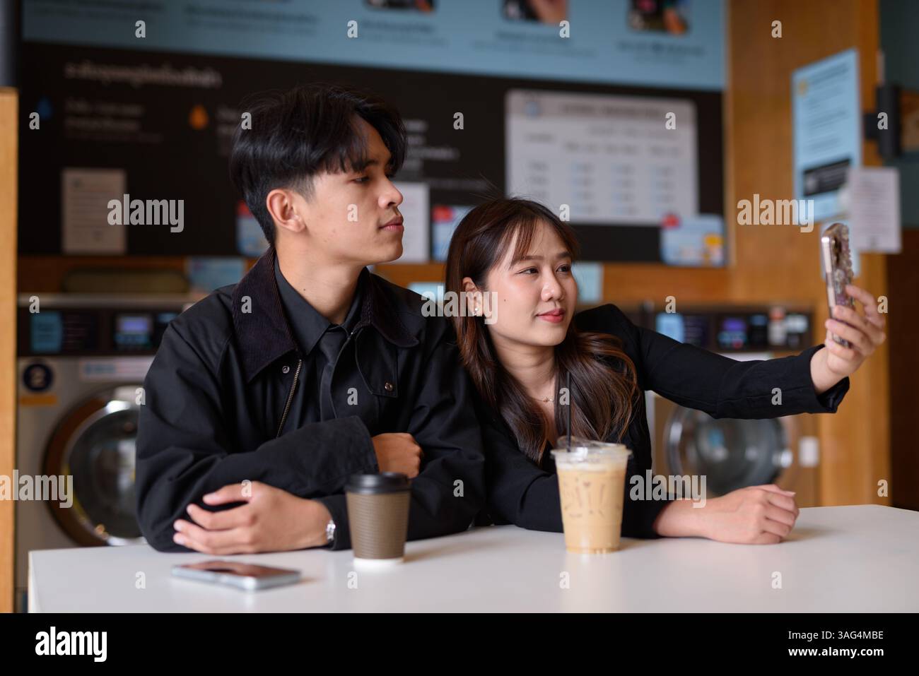 Giovane coppia asiatica felice seduta in lavanderia a bere caffè e a usare il telefono Foto Stock