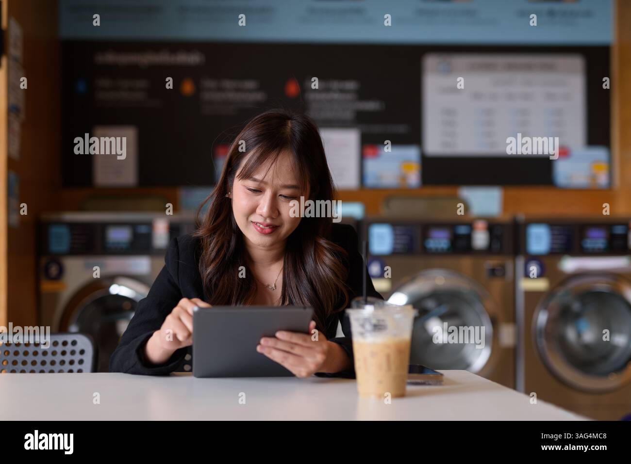 Donna seduta in lavanderia che usa un tablet digitale e beve un caffè ghiacciato Foto Stock