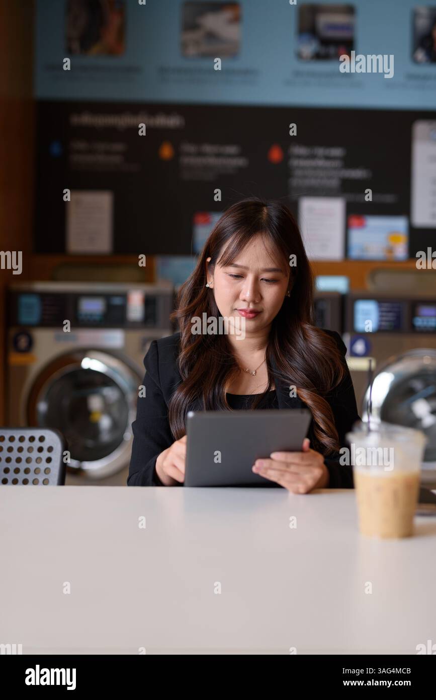 Donna seduta in lavanderia che usa un tablet digitale e beve un caffè ghiacciato Foto Stock