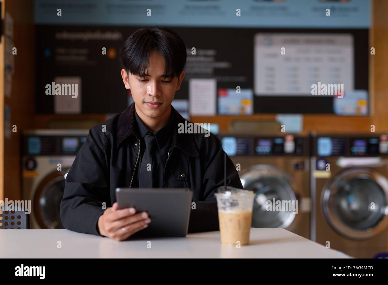 Uomo seduto in un negozio di lavanderia che usa un tablet digitale e beve un caffè Foto Stock