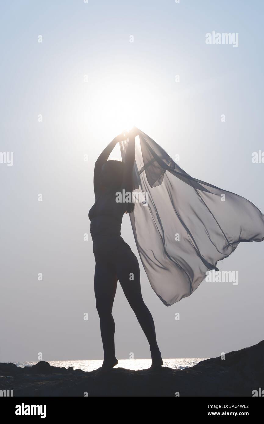 Una silhouette mozzafiato di una graziosa ballerina che regge un tessuto fluttuante sullo splendido tramonto sulla spiaggia Foto Stock