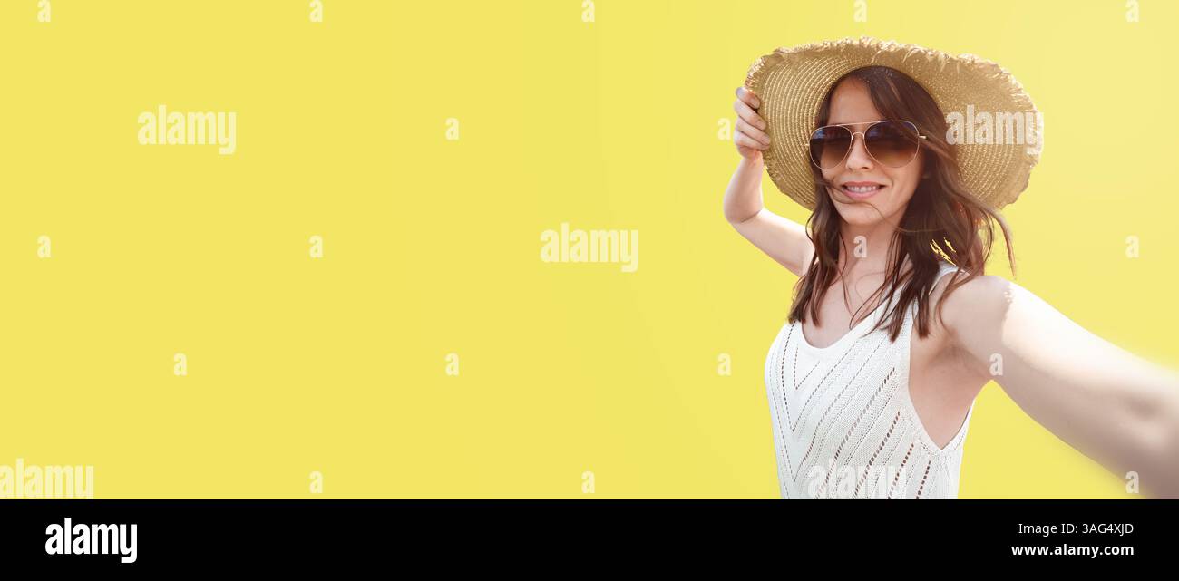 Una donna sorridente di 30-35 anni con un cappello a tesa larga e occhiali da sole su sfondo giallo, si fa un selfie. Un concetto per viaggi e blog, Foto Stock