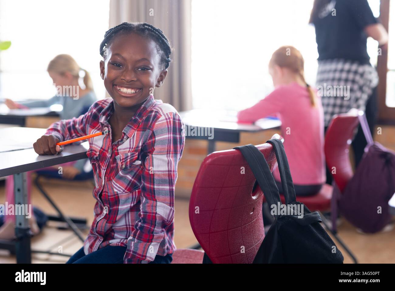 Sorridente ragazza afroamericana che tiene la matita in classe con altri studenti che studiano a scuola Foto Stock