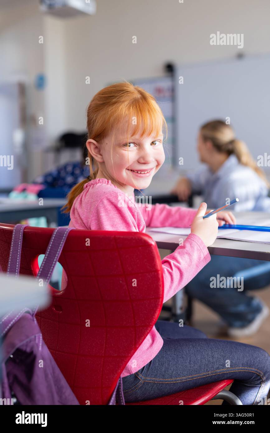 A scuola, la ragazza sorride e tiene in mano una matita mentre si siede alla scrivania in classe Foto Stock