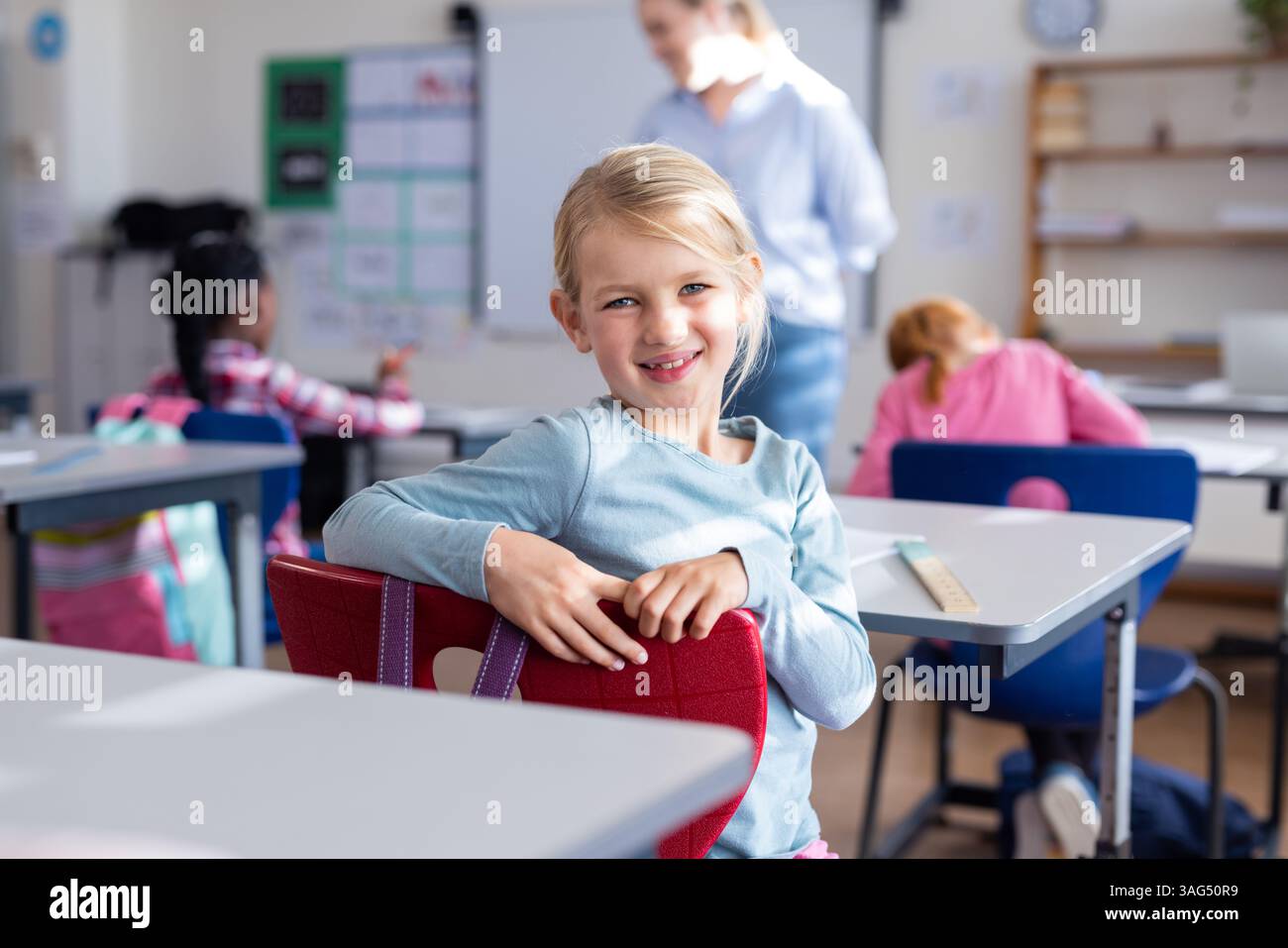 A scuola, una ragazza sorridente seduta alla scrivania in classe con i compagni di classe Foto Stock