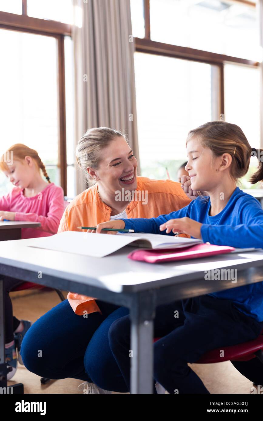 A scuola, una professoressa sorride e aiuta il ragazzo a leggere alla scrivania Foto Stock