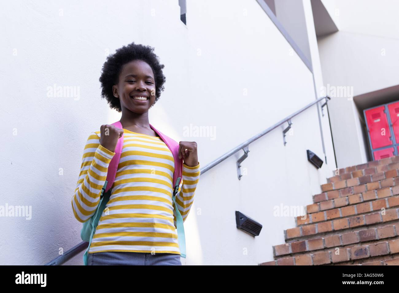 Ragazza afroamericana sorridente con zaino in piedi sulla scala della scuola, pronta per la lezione Foto Stock