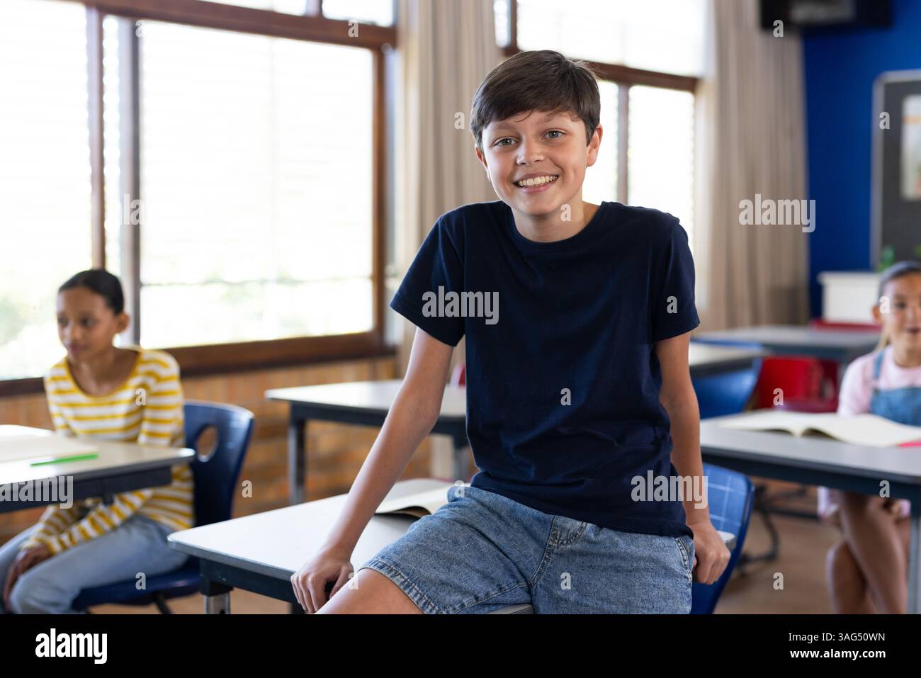 Ragazzo sorridente seduto sulla scrivania in classe con gli studenti in background a scuola Foto Stock