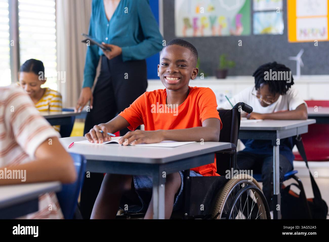 A scuola, ragazzo in sedia a rotelle sorride e scrive alla scrivania in classe Foto Stock