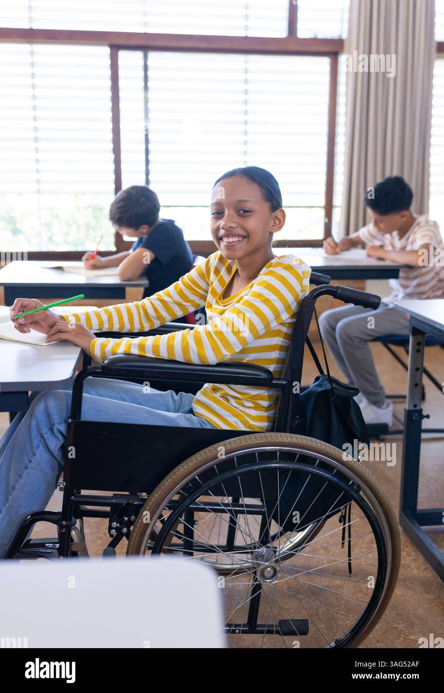 A scuola, ragazza in sedia a rotelle sorride e scrive in classe con i compagni di classe Foto Stock