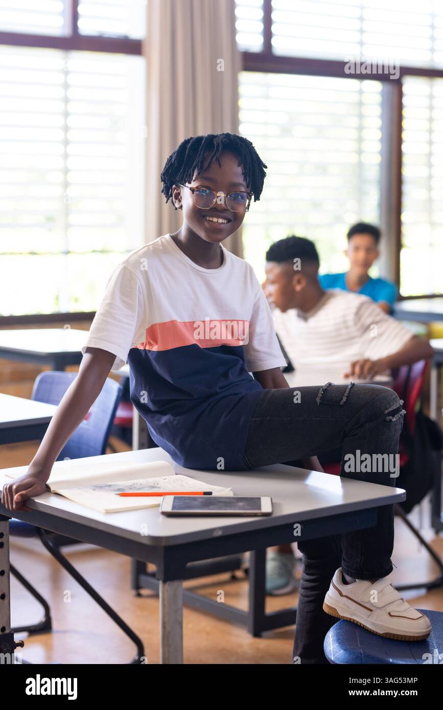 A scuola, uno studente sorridente seduto sulla scrivania con un tablet e un notebook nelle vicinanze Foto Stock