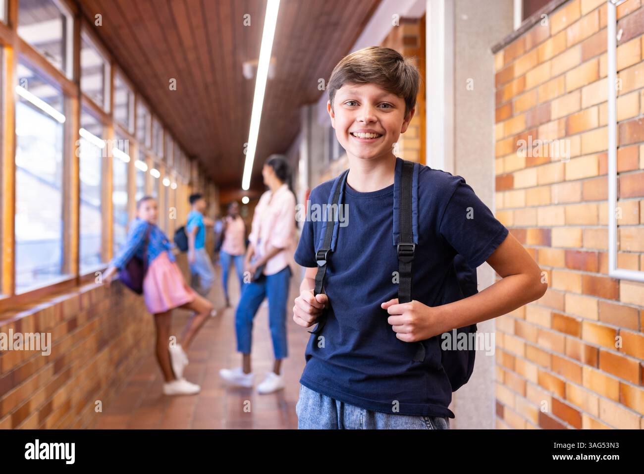 Ragazzo sorridente con zaino in piedi nel corridoio della scuola con compagni di classe sullo sfondo Foto Stock