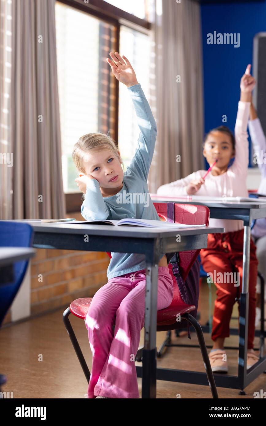 A scuola, ragazza che alza la mano mentre siede alla scrivania in classe Foto Stock