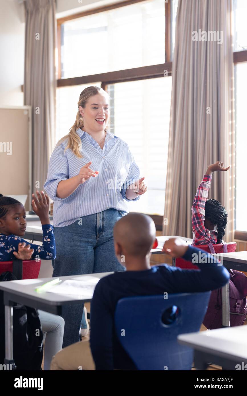 A scuola, un'insegnante donna che si impegna con studenti diversi alzando la mano nella discussione in classe Foto Stock