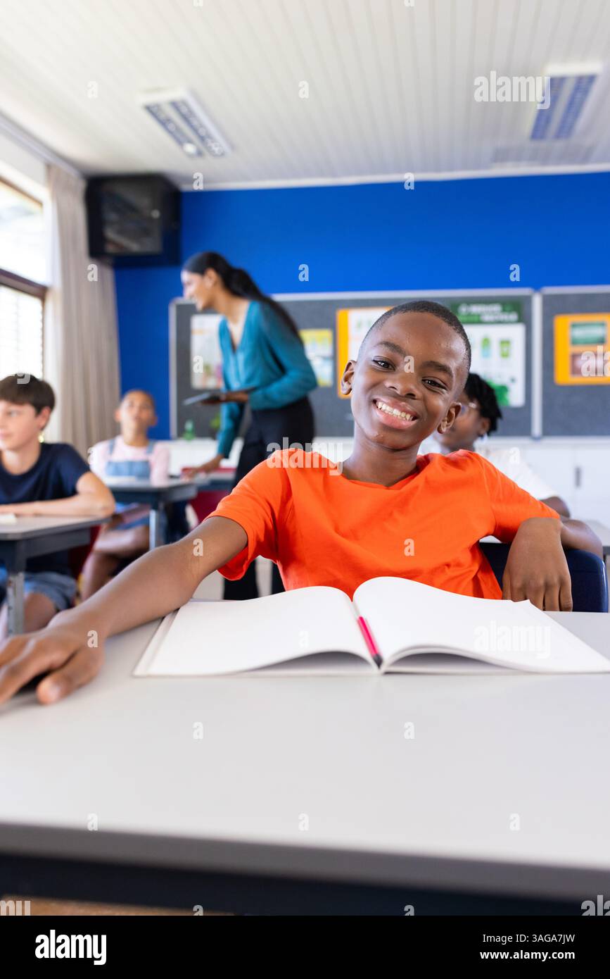 Ragazzo sorridente in classe con notebook aperto, circondato da compagni di classe a scuola Foto Stock
