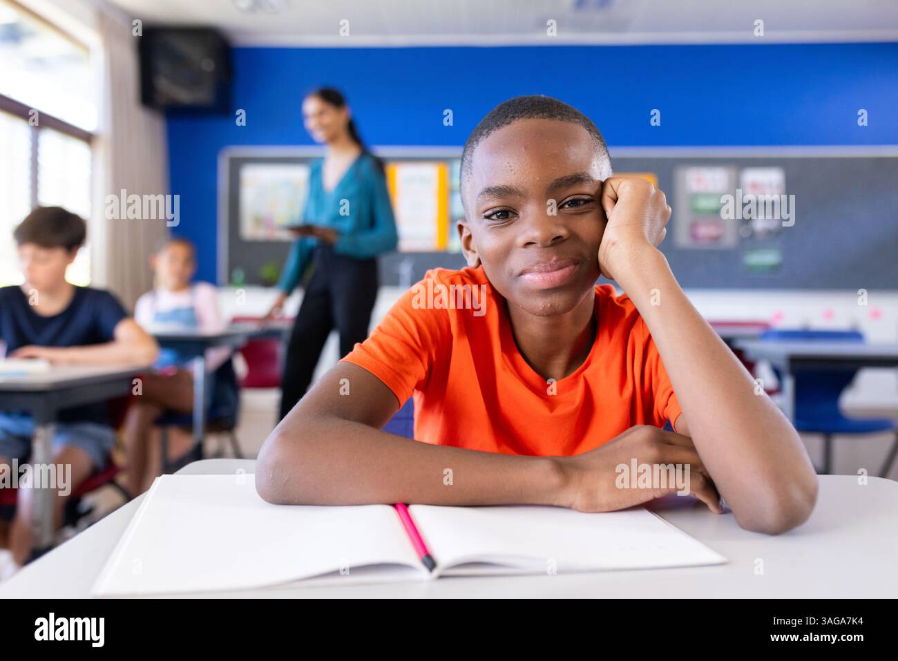 A scuola, ragazzo seduto alla scrivania con un notebook aperto, sorridendo alla macchina fotografica Foto Stock