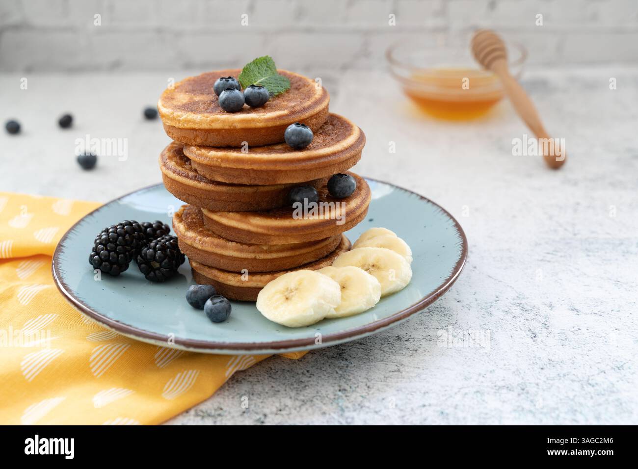 Frittelle dolci e soffici sovrapposte adornate con banana e frutti di bosco. Con il miele su piatti blu su un piano da tavolo grigio chiaro con un panno giallo Foto Stock
