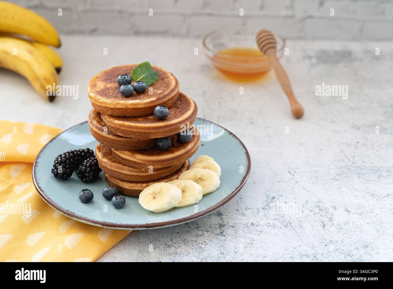 Frittelle dolci e soffici sovrapposte adornate con banana e frutti di bosco. Con il miele su piatti blu su un piano da tavolo grigio chiaro con un panno giallo Foto Stock