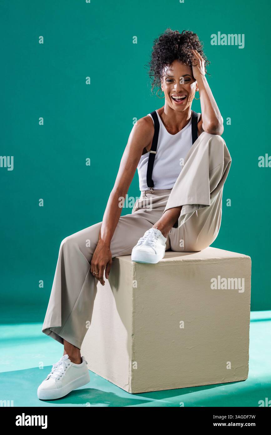 Donna sorridente seduta su un cubo che indossa un outfit casual in un moderno studio. Moda, modello, stile, ritratto, contemporaneo, sicurezza Foto Stock