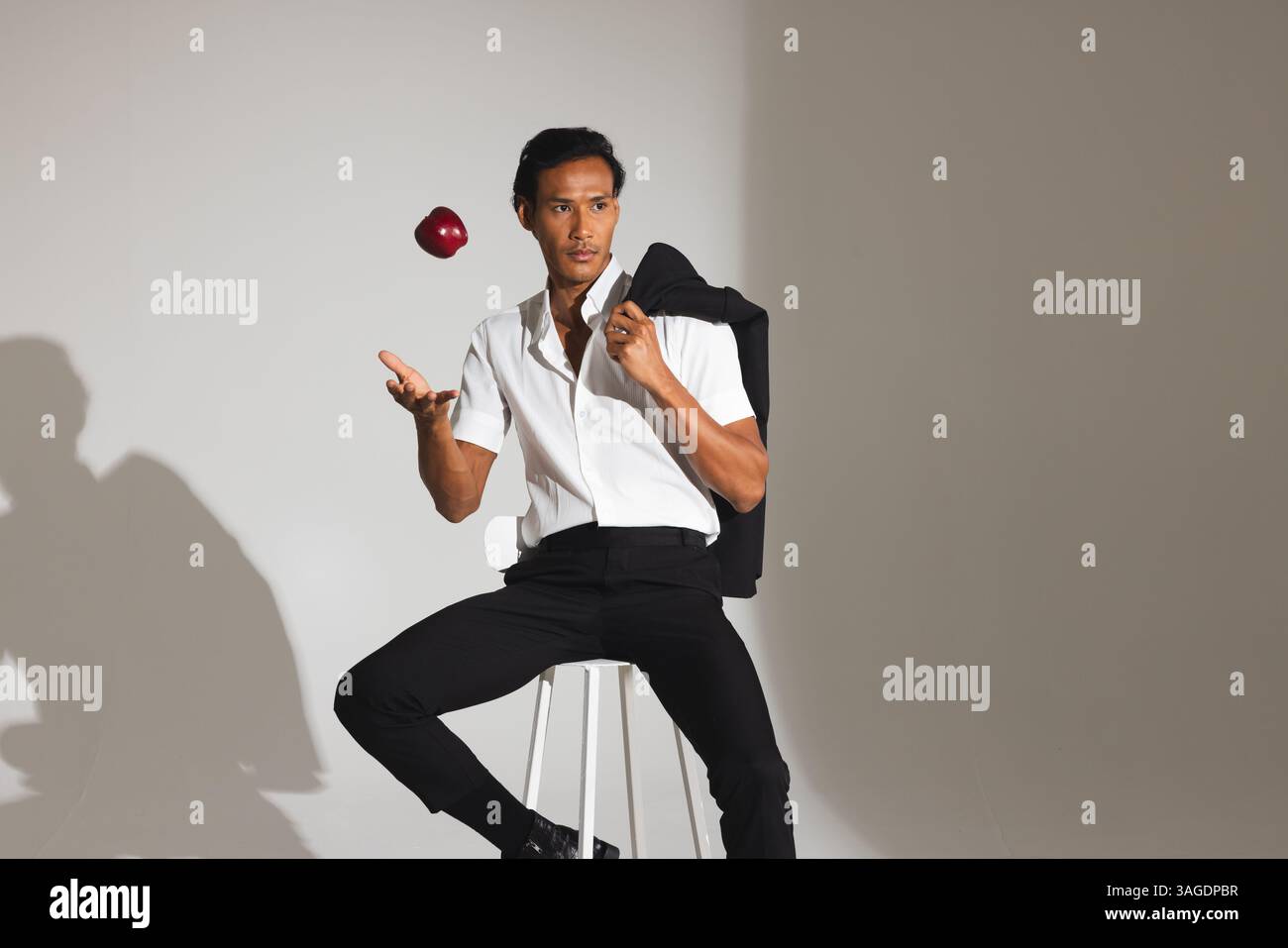 Uomo elegante in camicia bianca che lancia la mela seduto sullo sgabello in studio. Moda, minimalismo, casual, trendy, eleganza, in posa Foto Stock