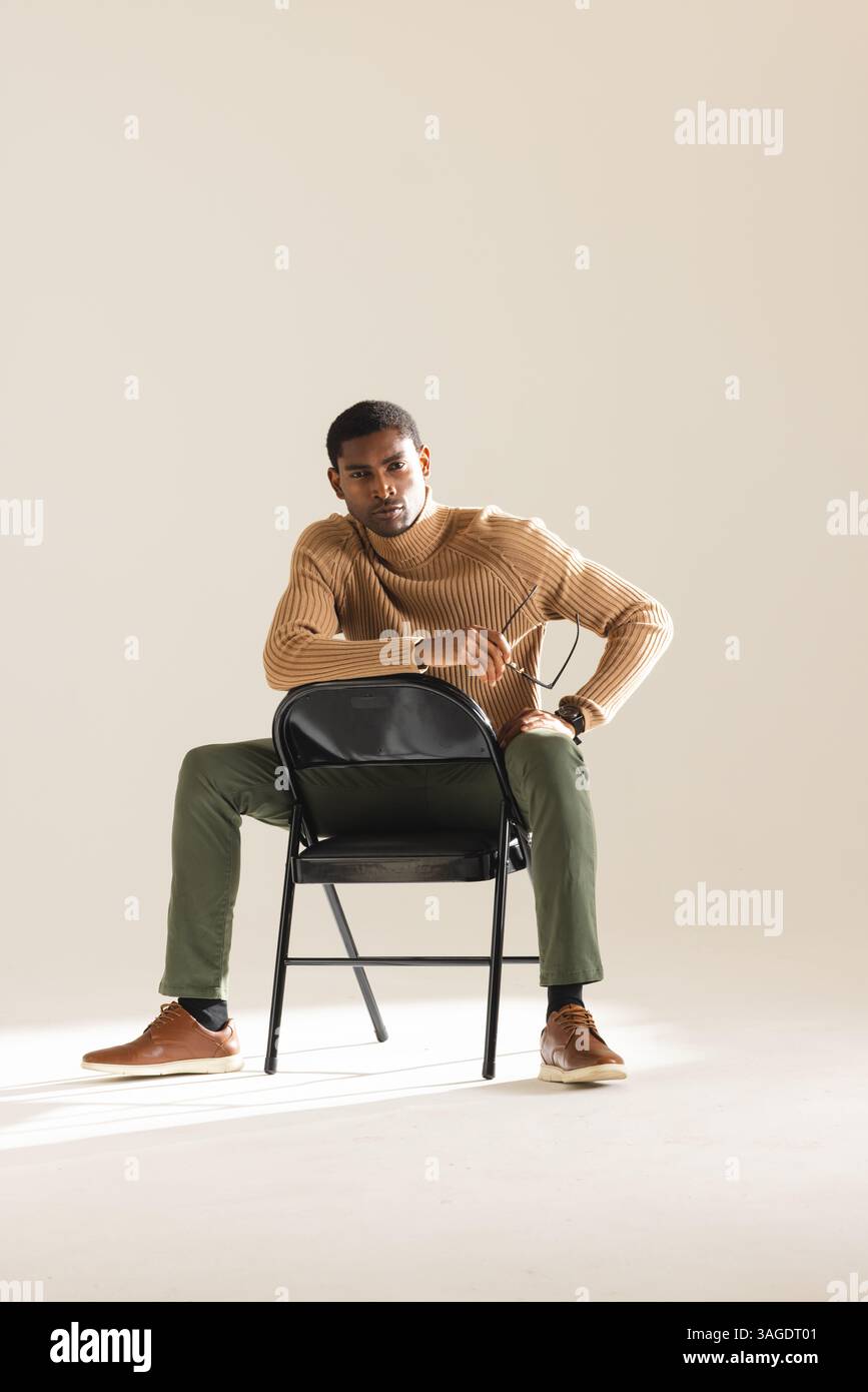 Uomo afro-americano in un abito elegante seduto con sicurezza su una sedia in studio. Moda, sicurezza, stile, individuale, ritratto, modello Foto Stock