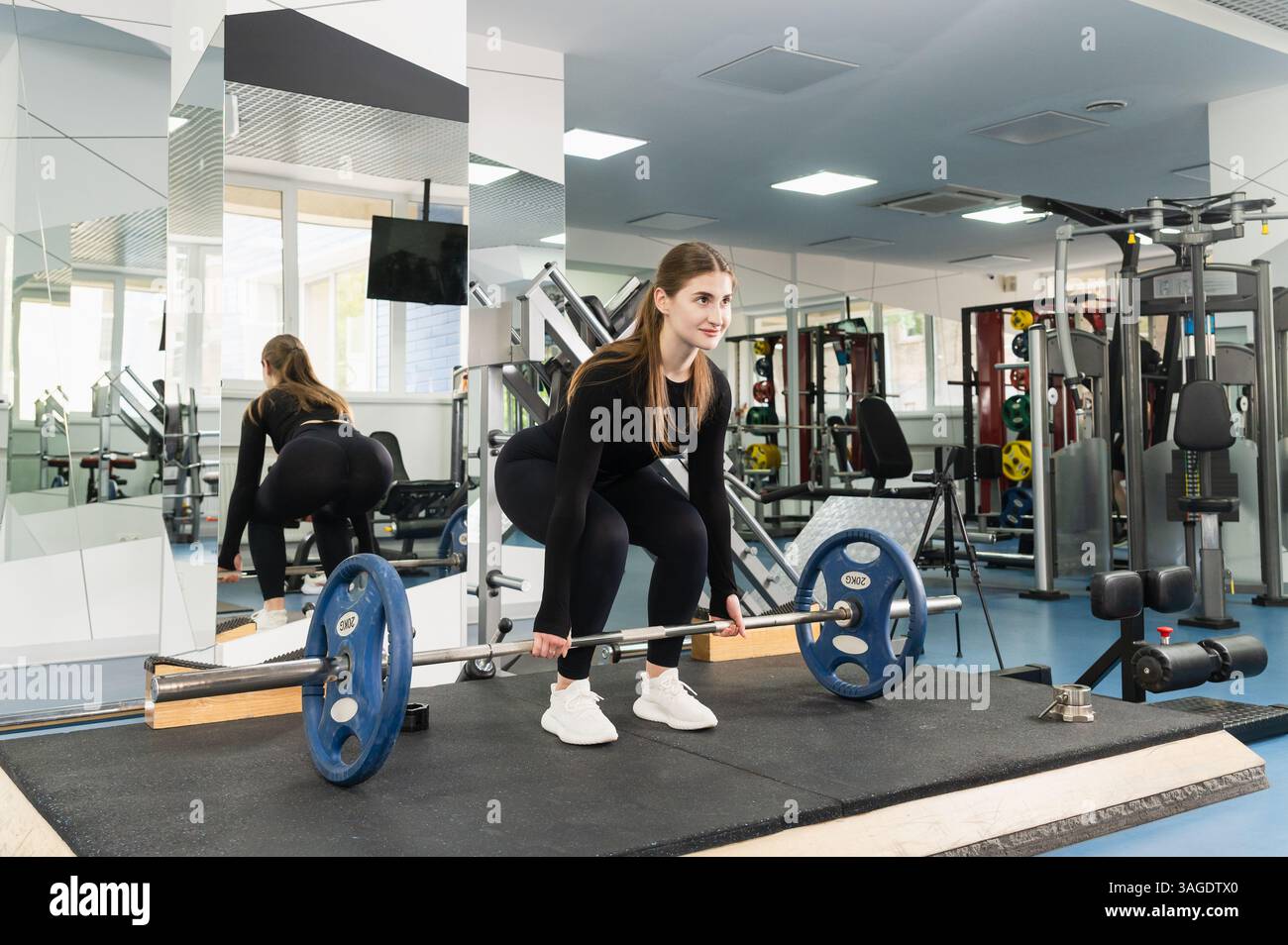 Vesti la giovane donna con un abito nero e preparati a sollevare un barbell con piatti blu in una palestra moderna e spaziosa. Vista laterale e posteriore, specchio e attrezzatura Foto Stock Vesti la giovane donna con un abito nero e preparati a sollevare un barbell con piatti blu in una palestra moderna e spaziosa. Vista laterale e posteriore, specchio e attrezzatura Foto Stock