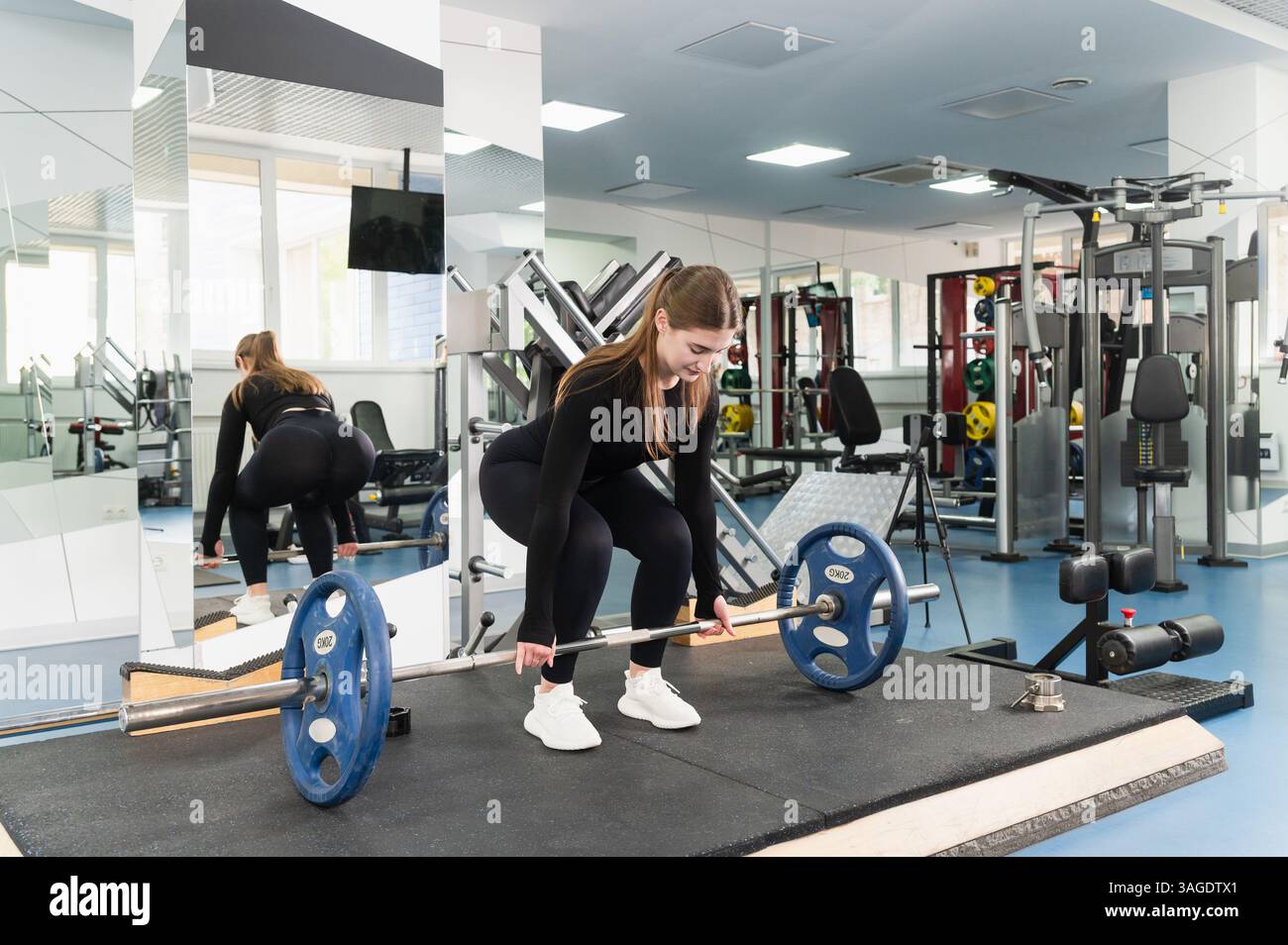 Vesti la giovane donna con un abito nero e preparati a sollevare un barbell con piatti blu in una palestra moderna e spaziosa. Vista laterale e posteriore, specchio e attrezzatura Foto Stock Vesti la giovane donna con un abito nero e preparati a sollevare un barbell con piatti blu in una palestra moderna e spaziosa. Vista laterale e posteriore, specchio e attrezzatura Foto Stock