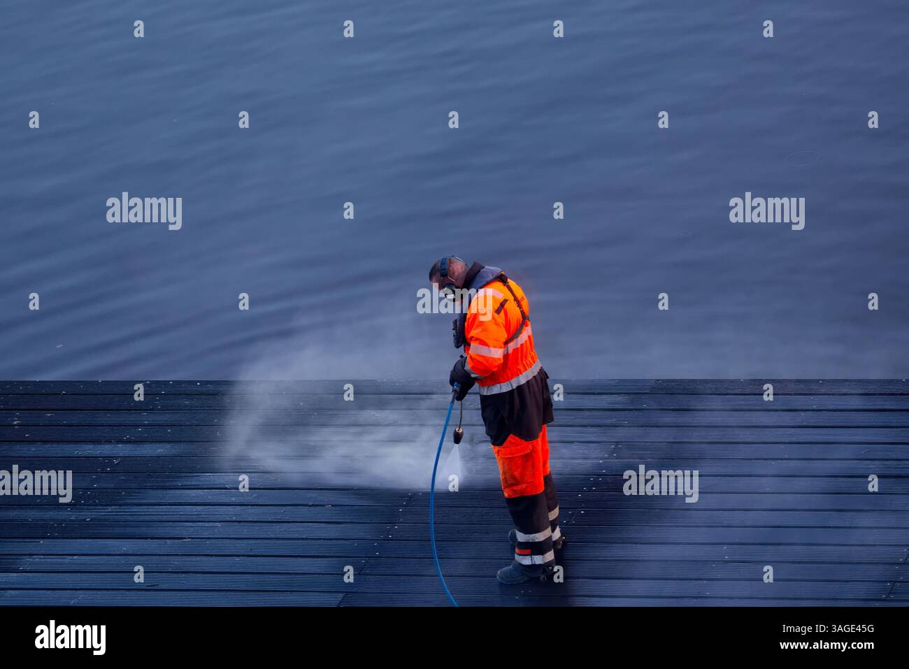Un lavoratore con attrezzatura di sicurezza arancione e nera utilizza un tubo dell'acqua ad alta pressione per pulire un molo di legno nelle acque calme di Amsterdam, Paesi Bassi. Foto Stock