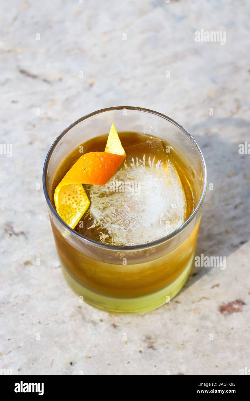Sopra la vista di un cocktail di whisky con un grande cubetto di ghiaccio decorato con una scorza d'arancia sopra un tavolo in marmo bege Foto Stock