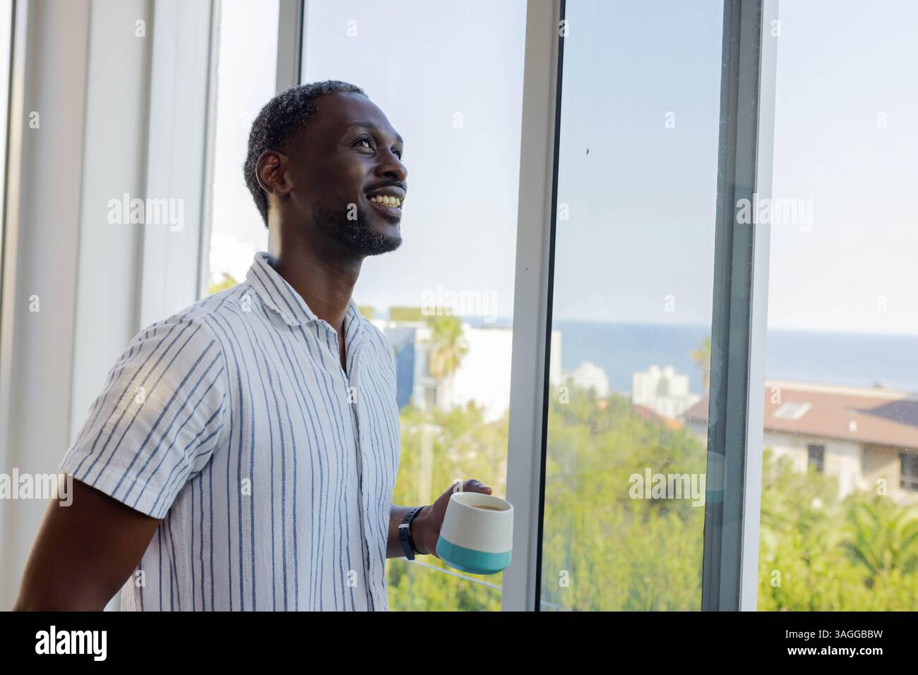 Sorridente uomo afroamericano che si gusta il caffè mattutino dalla finestra, con vista panoramica. Relax, stile di vita, bevande, contemplativo, tranquillità, Foto Stock