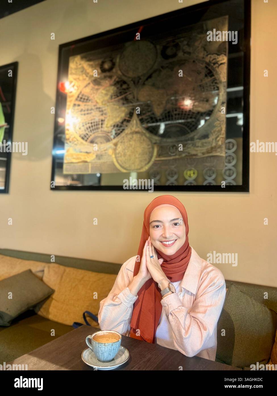 Sorridente giovane donna in un velo che si gusta un caffè al tavolo di un bar. Luce naturale, tonalità calde, atmosfera rilassata, ambiente interno. Foto Stock