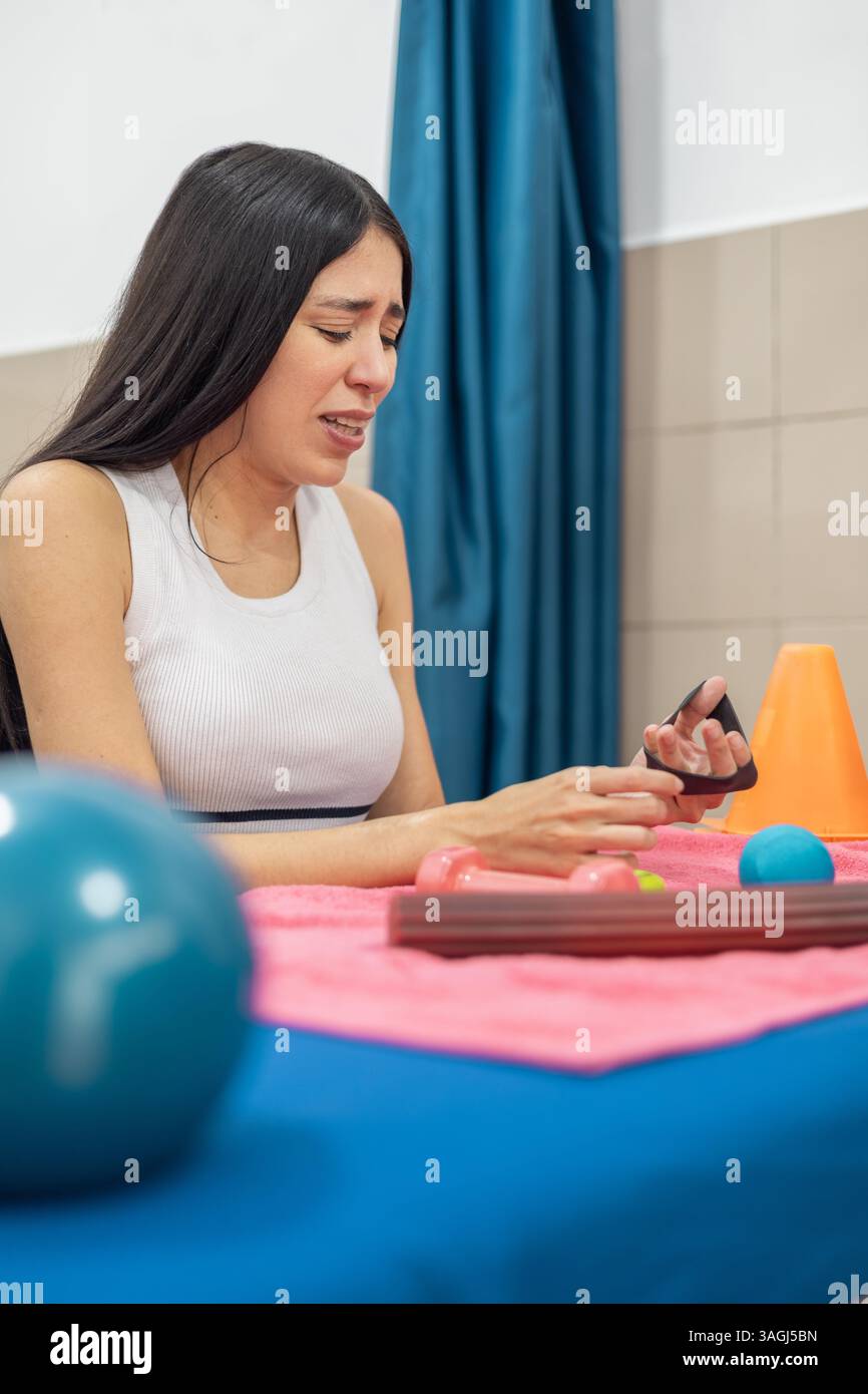 Una donna guarda la sua mano dopo aver completato una sessione di riabilitazione con vari attrezzi su un tavolo coperto da un asciugamano rosa. Foto Stock