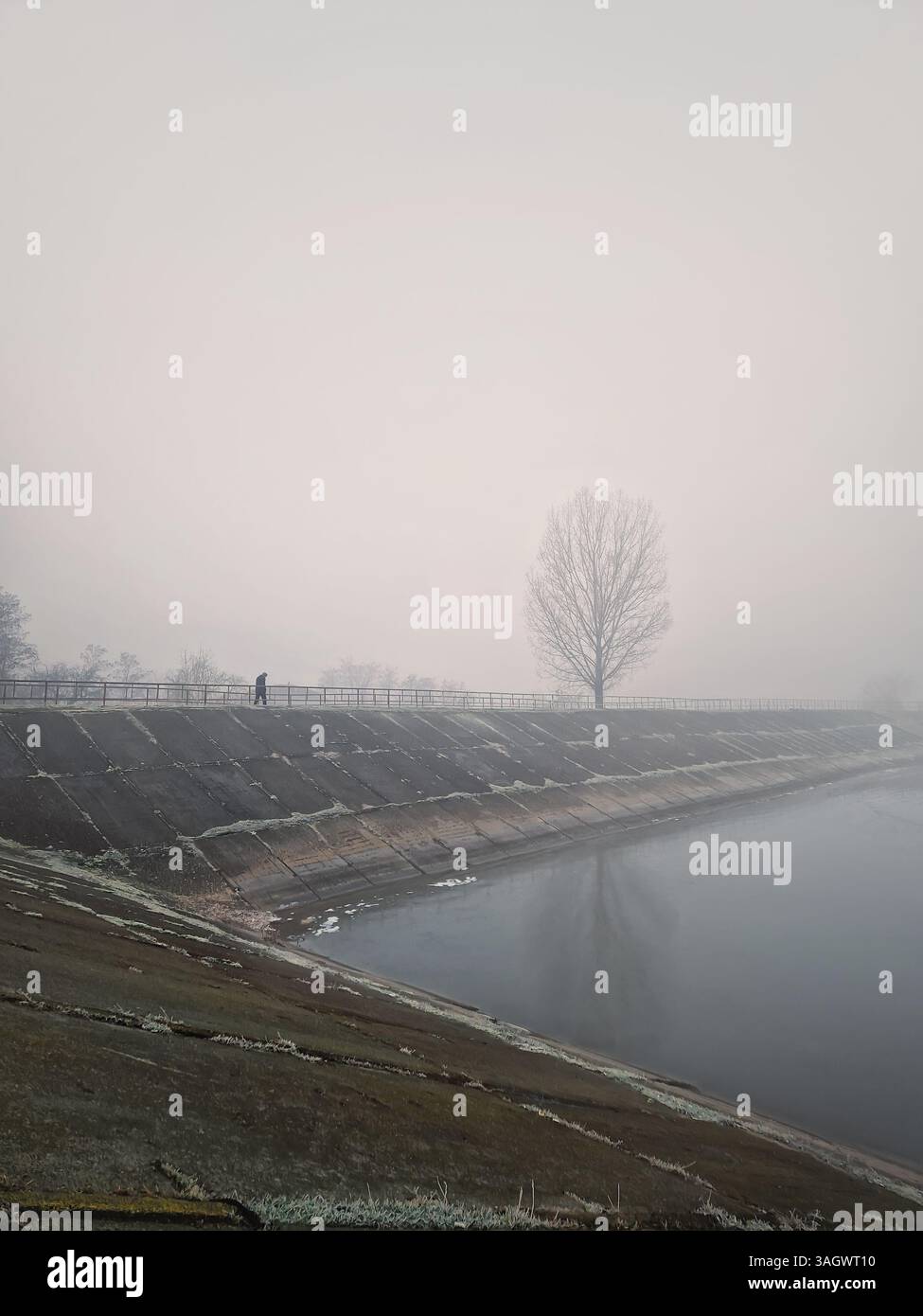 Fitta nebbia nella mattina d'inverno vicino al lago, con una persona che cammina sulla diga d'acqua attraverso la fitta nebbia - Immagine stock catturata con smartphone