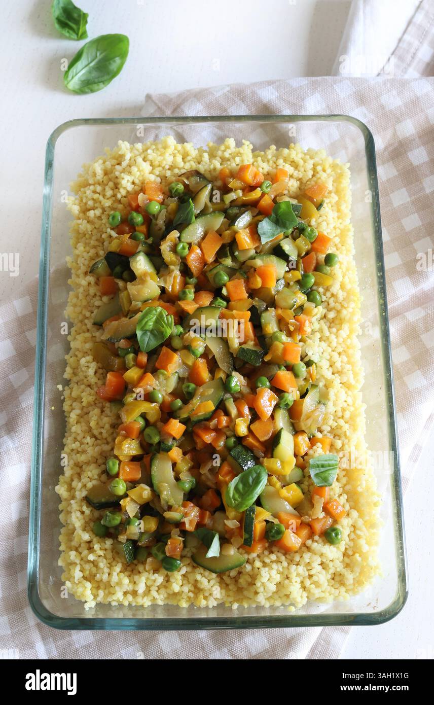 Couscous con verdure (zucchine, peperoni, carote, erbe) in un vassoio di vetro isolato su un tavolo di legno bianco. Cibo sano e vegetariano. Foto Stock