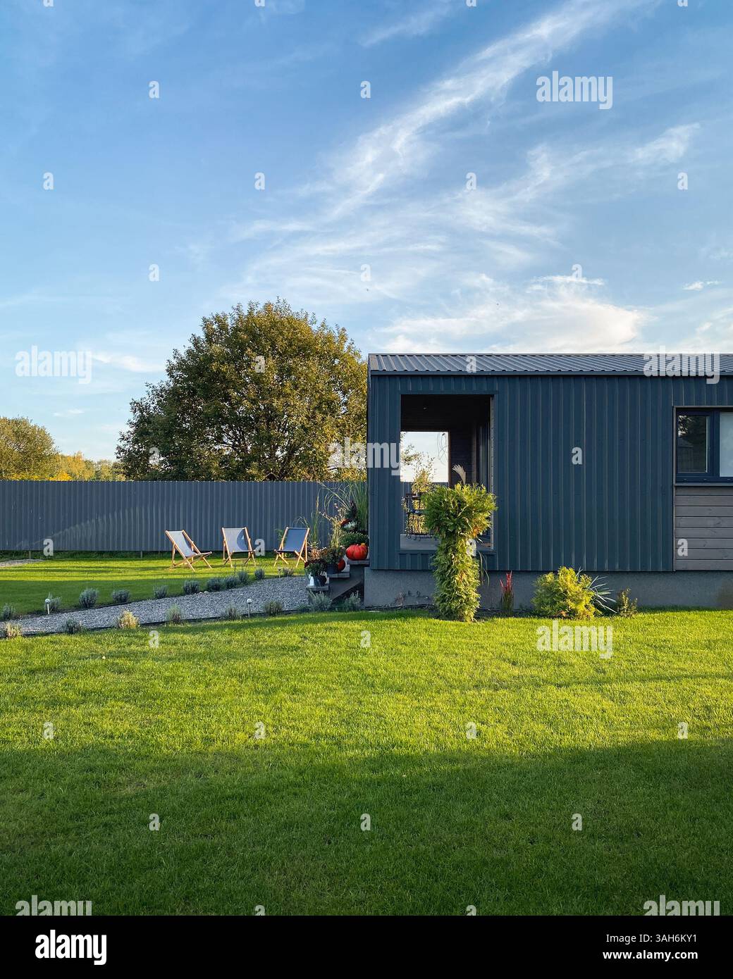 Cortile posteriore con i raggi del sole pomeridiano: Tre sdraio su un prato, portico della casa estiva e sentiero pedonale. diversi alberi dietro la recinzione grigia. Foto Stock