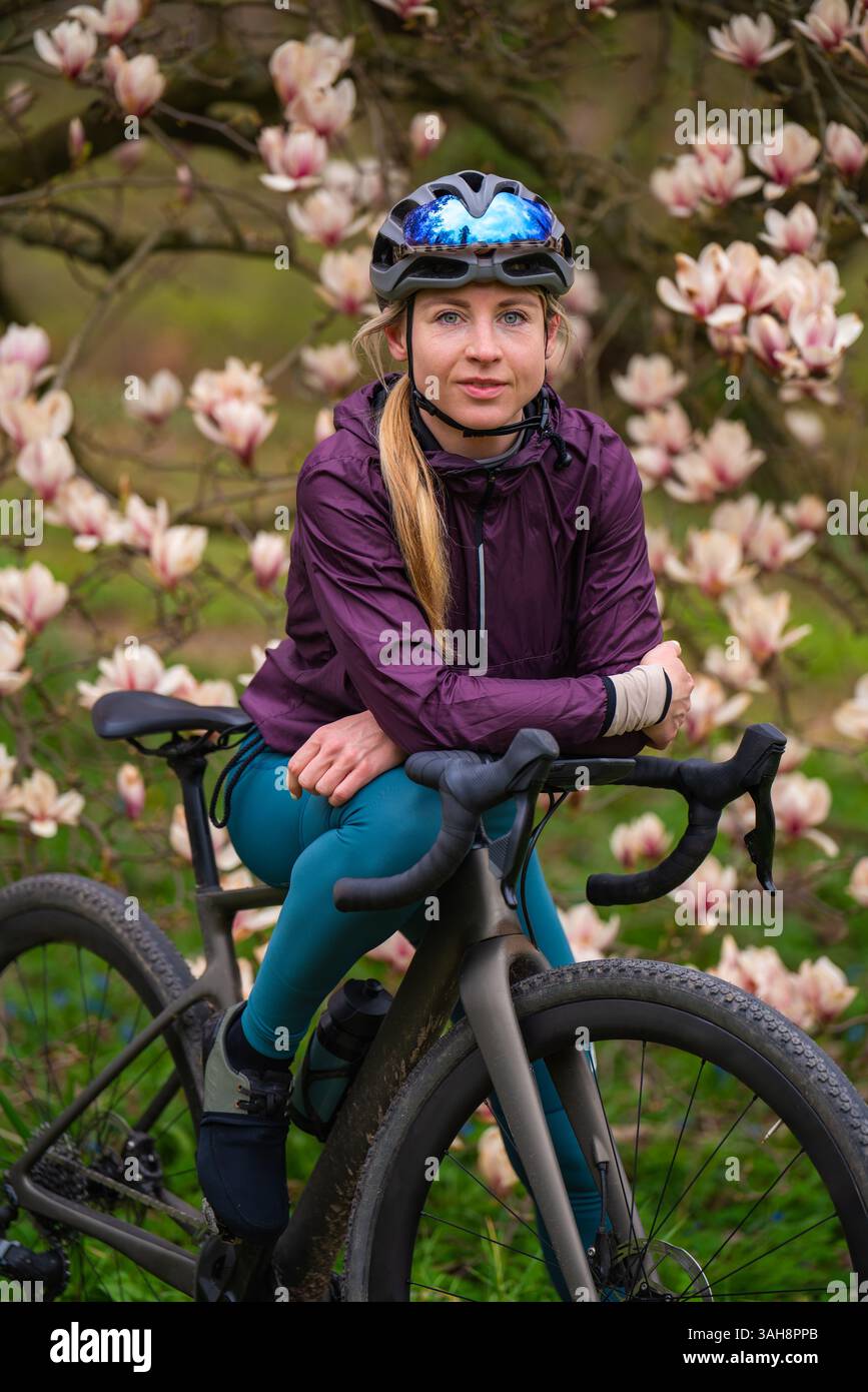Ciclista donna in uniforme sportiva, casco in piedi con Gravel bike vicino alla fioritura dell'albero di magnolia. Primavera, combinazione di sport e bellezza naturale. Foto Stock