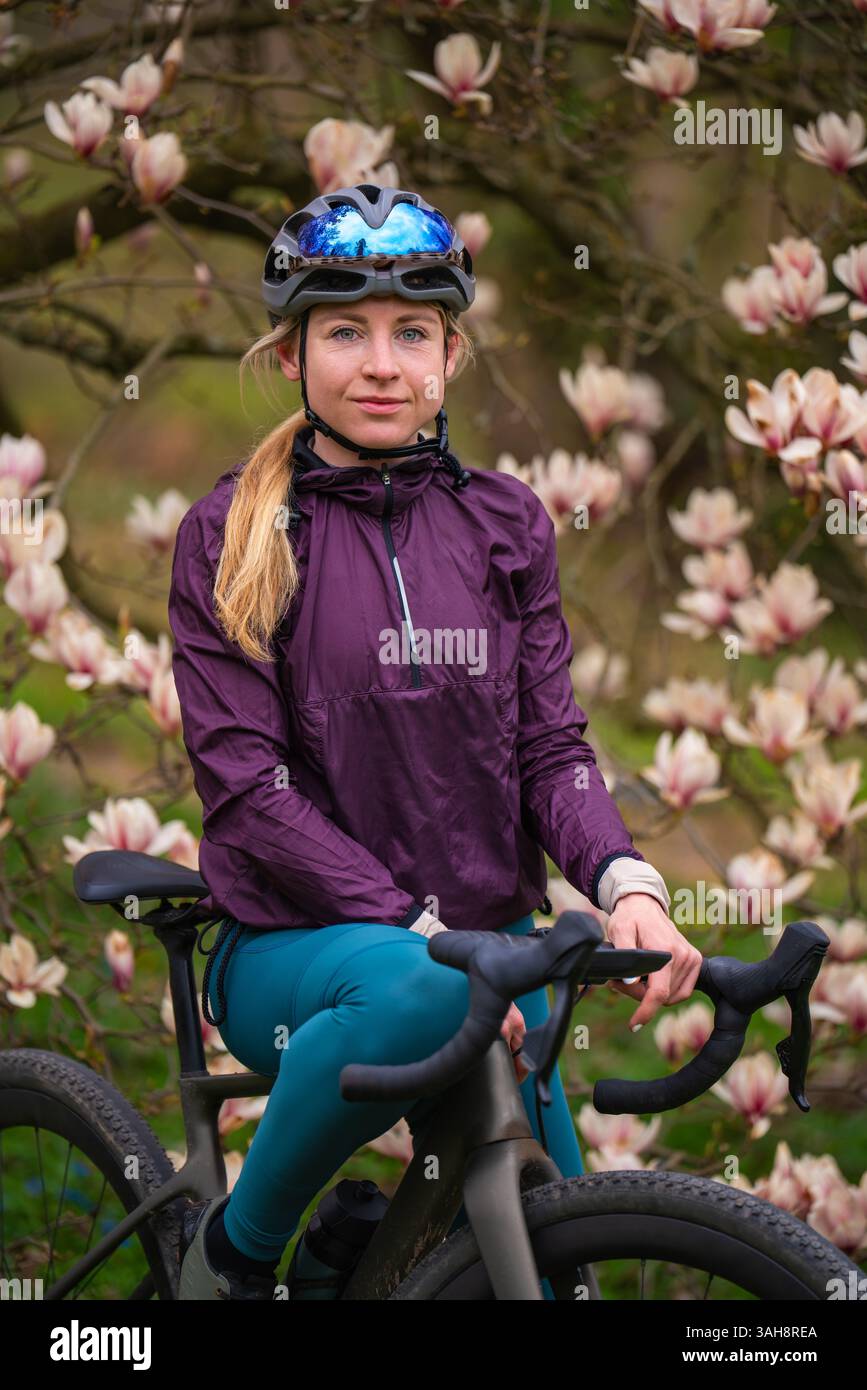 Ciclista donna in uniforme sportiva, casco in piedi con Gravel bike vicino alla fioritura dell'albero di magnolia. Primavera, combinazione di sport e bellezza naturale. Foto Stock