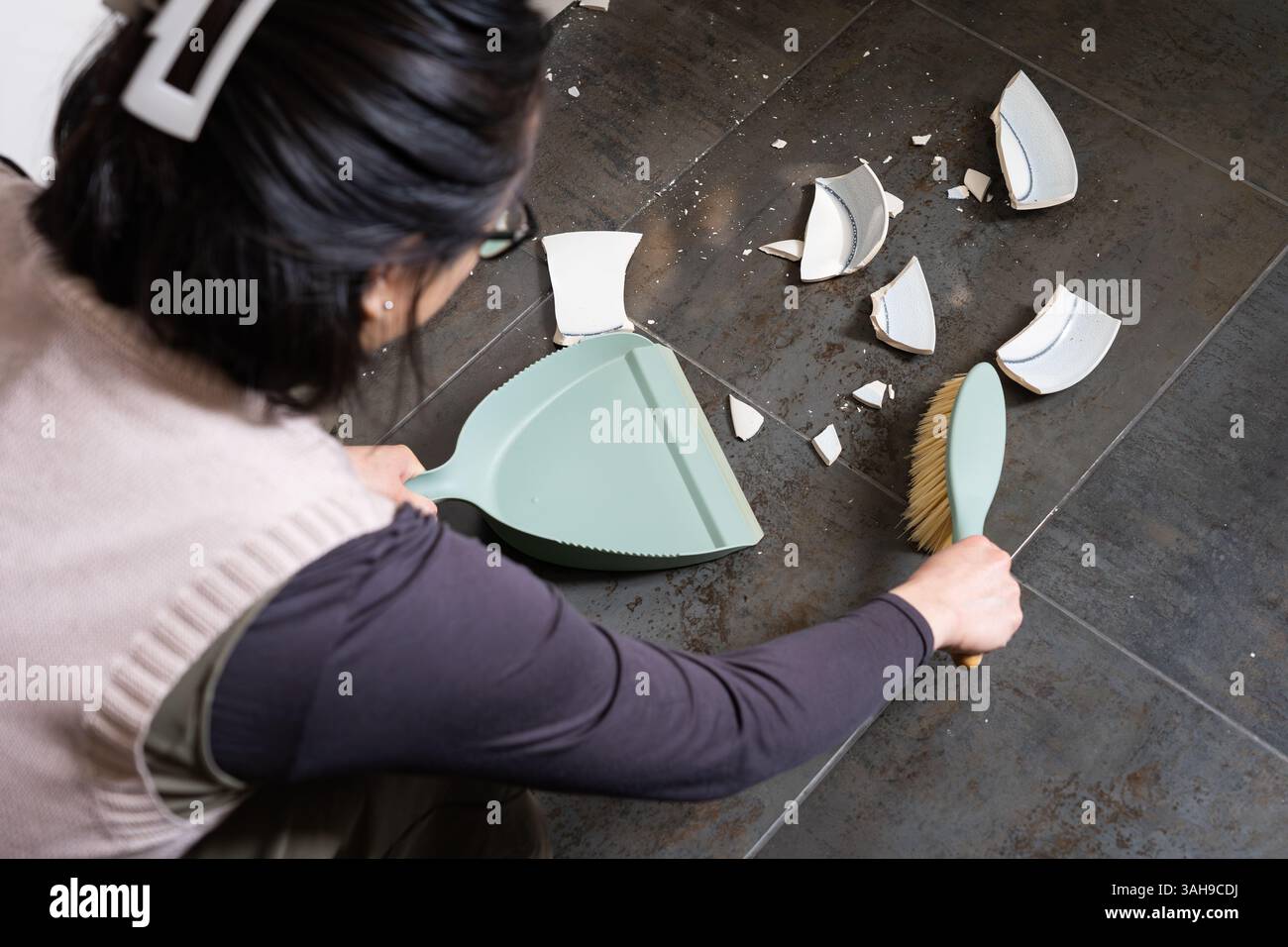 Una donna che spazza via un piatto rotto con una padella e una spazzola dal pavimento di una cucina, in Inghilterra. Concetto: Broken china, lavori domestici, goffa, goffa Foto Stock