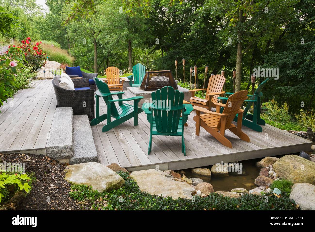Mobili in vimini marroni, sedie Adirondack in legno verde e marrone su un terrazzo in legno grigio con focolare in un giardino in pendenza in estate. Foto Stock