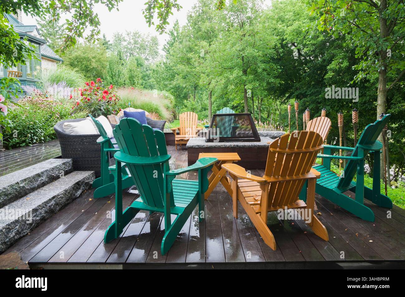 Mobili in vimini marroni, sedie Adirondack in legno verde e marrone su un terrazzo in legno grigio con focolare in un giardino in pendenza in estate. Foto Stock