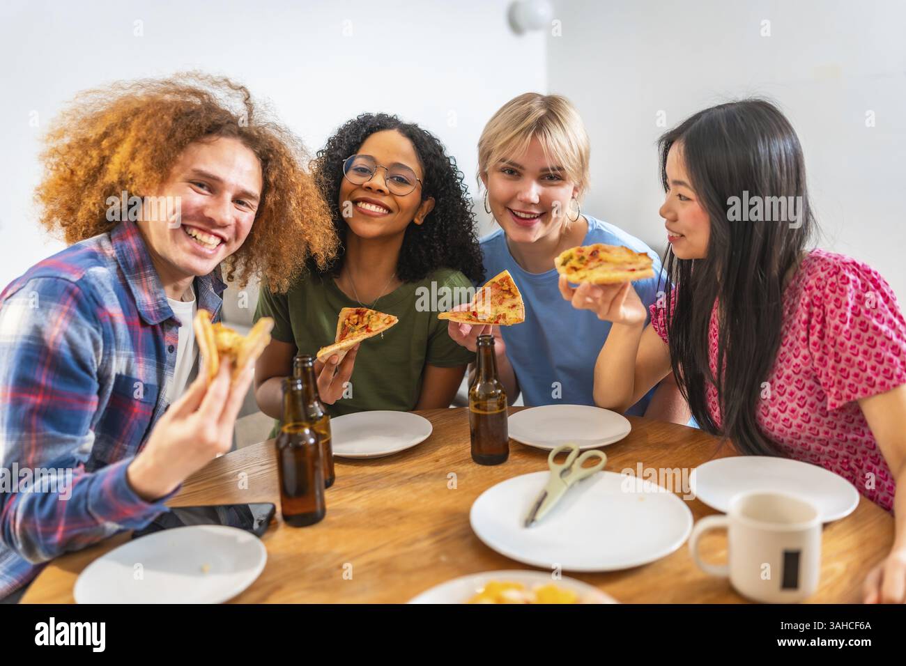 Quattro giovani e allegri amici di diverse origini gustando pizza e birra insieme a casa, celebrando l'amicizia e i bei momenti Foto Stock