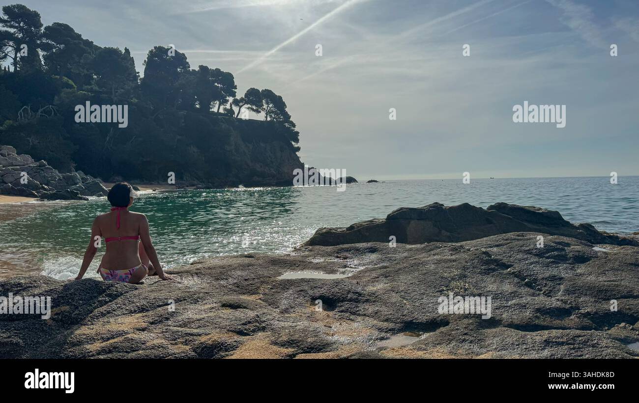 Una donna in bikini rosa si siede sulla spiaggia sabbiosa di Lloret de Mar, Spagna. Il suo volto non è visibile, concentrandosi sull'atmosfera rilassata della spiaggia e. Foto Stock