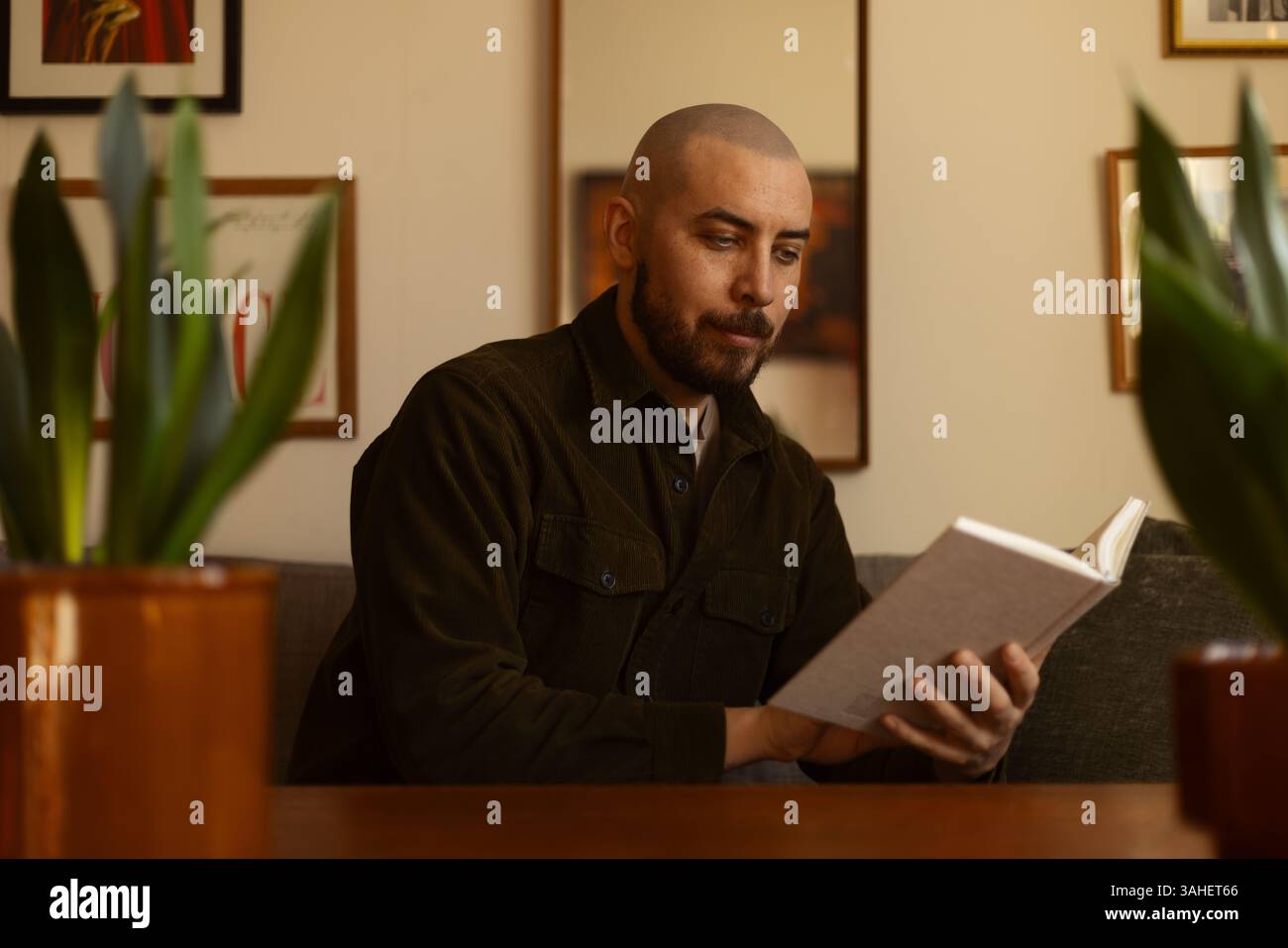 Ritratto di un giovane uomo caucasico con la barba che scrive in un libro seduto in un accogliente soggiorno con una tazza di caffè da leggere. Foto Stock