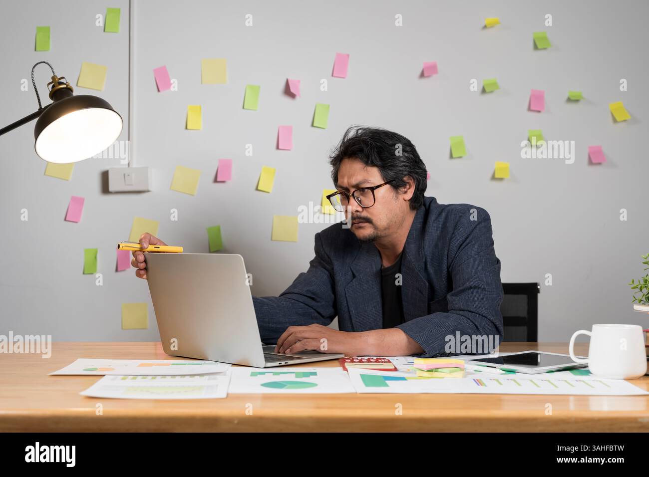 Un uomo professionista che indossa gli occhiali sta lavorando intensamente su un notebook in un ambiente accogliente, che simboleggia la dedizione e la ricerca intellettuale. Un uomo con i capelli disordinati Foto Stock