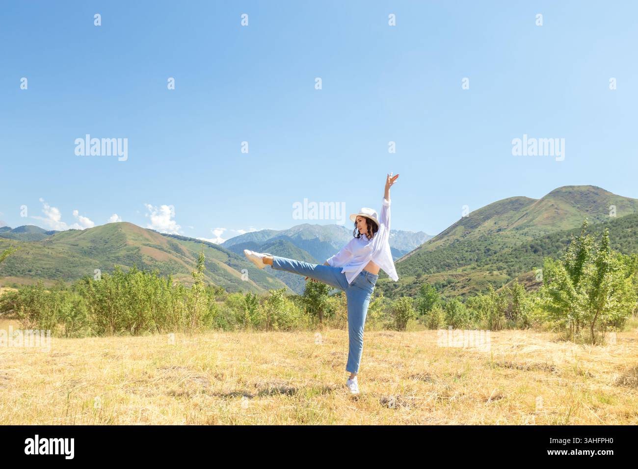 Una giovane donna allegra balla liberamente in un prato dorato circondato da alberi, con montagne sullo sfondo. Concetti di libertà, felicità, natura, Foto Stock