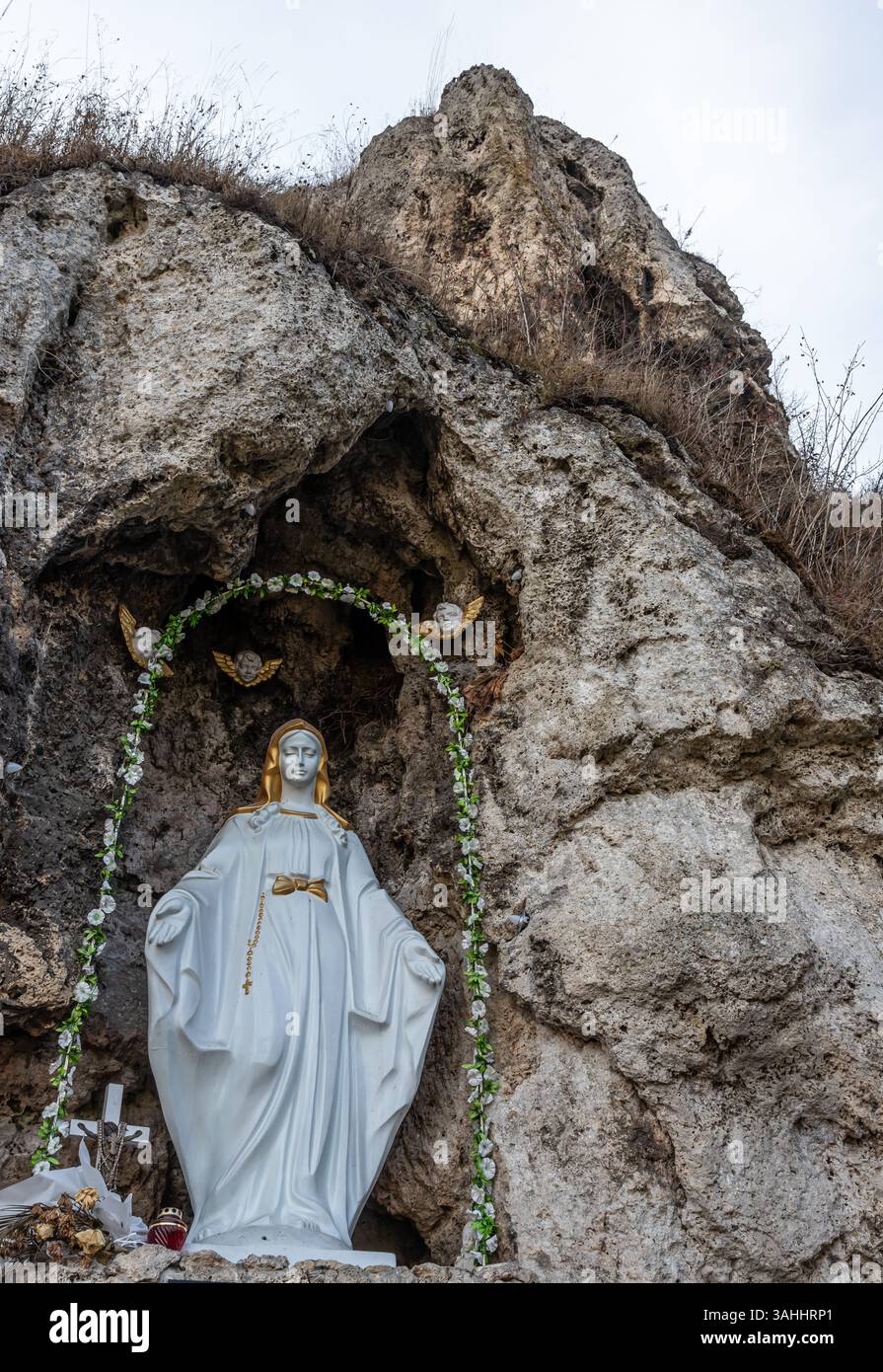 Una statua in marmo di una donna vestita di bianco sorge all'ingresso di una grotta rocciosa, adornata di fiori e farfalle, trasuda tranquillità in natura Foto Stock