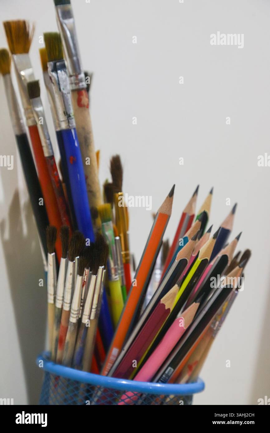 Una varietà di materiali d'arte sono disposti in una tazza di metallo, mostrando matite e pennelli colorati vivaci contro una semplice parete bianca. Foto Stock