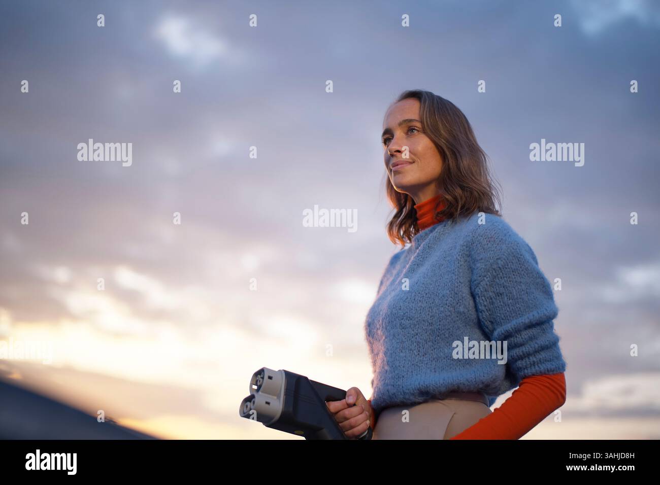 Una donna con un maglione blu regge un caricabatterie EV, in piedi contro un cielo nuvoloso al tramonto. Foto Stock