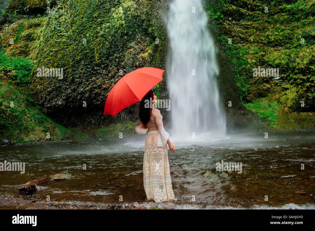 Donna con ombrello rosso in piedi vicino a una cascata circondata da lussureggianti foglie verdi. Silver Falls, Oregon, Stati Uniti Foto Stock