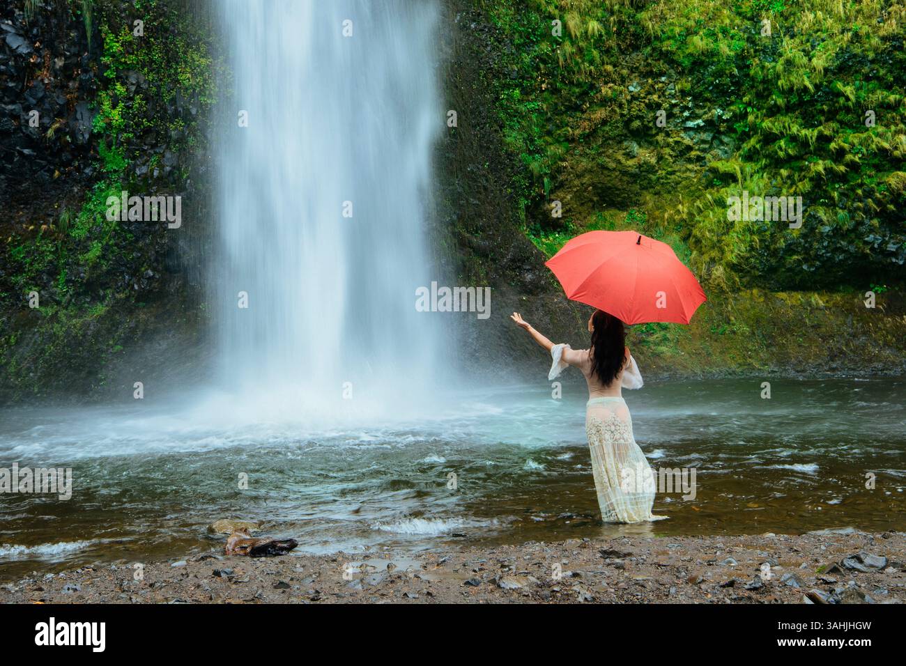 Donna con ombrello rosso si erge accanto a una cascata circondata da lussureggiante vegetazione. Silver Falls, Oregon, Stati Uniti Foto Stock